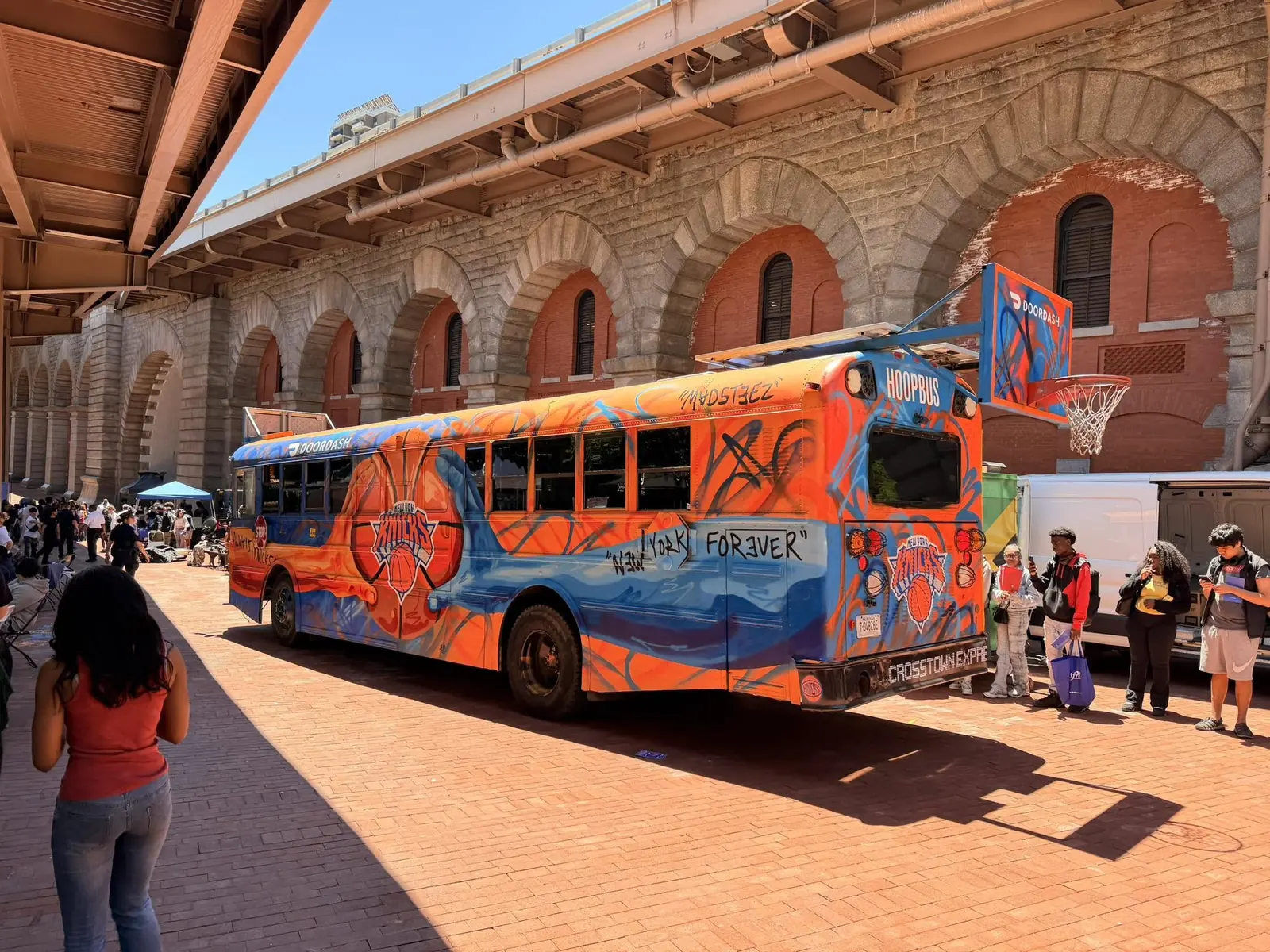 The Hoopbus parked under brick arches in the city, with a backboard extended over the pavement and a crowd gathered nearby