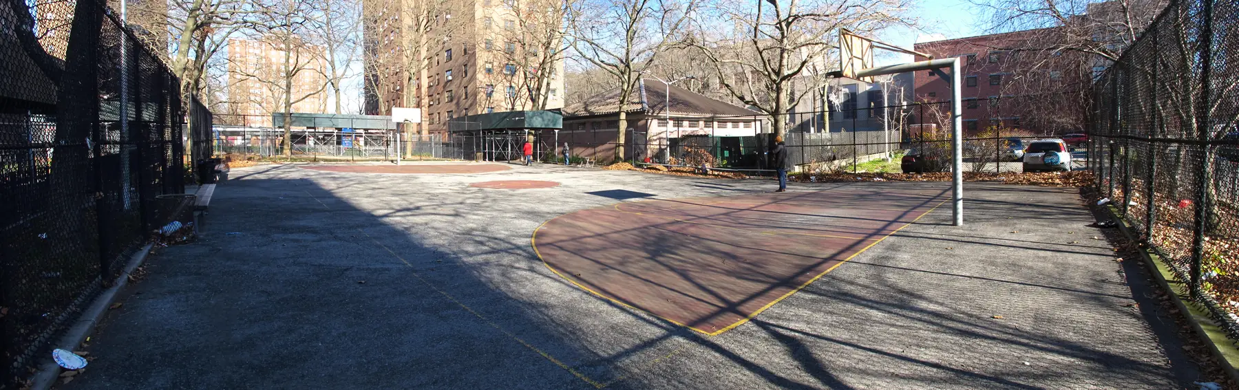 A wide view of the worn court at McKinley Houses, grey asphalt with faded red and yellow markings and bare trees overhead