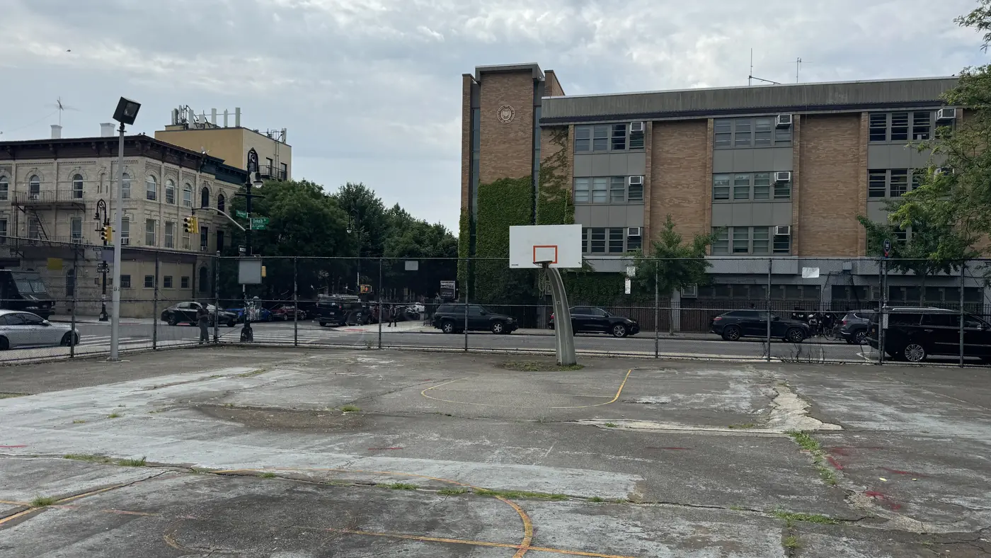 The far end of the Roosevelt Houses court, patched grey concrete with a single backboard and a faint yellow key line