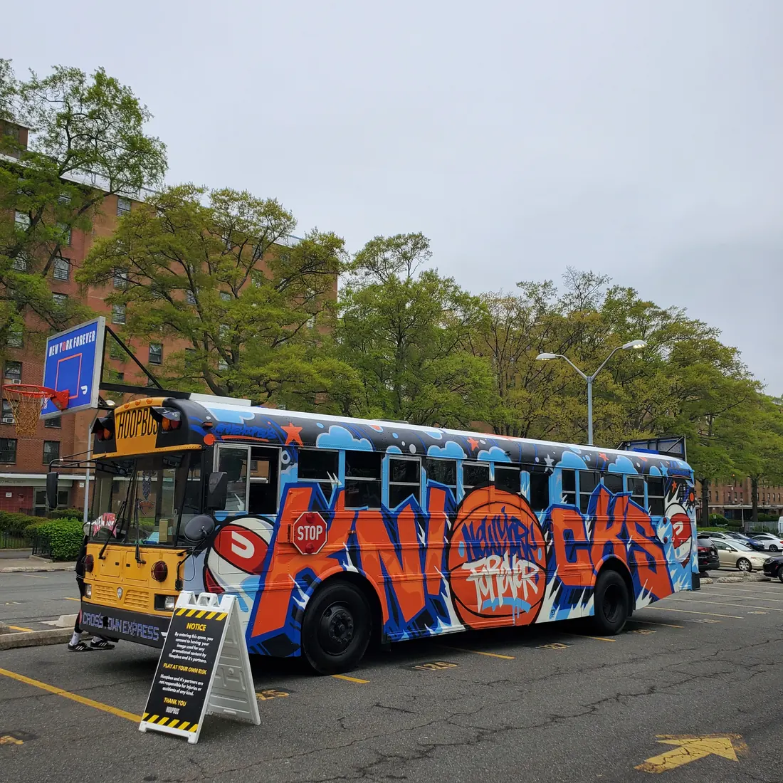 The Hoopbus, a school bus painted with a Knicks graffiti mural, parked beside a backboard reading New York Forever at Stapleton Houses